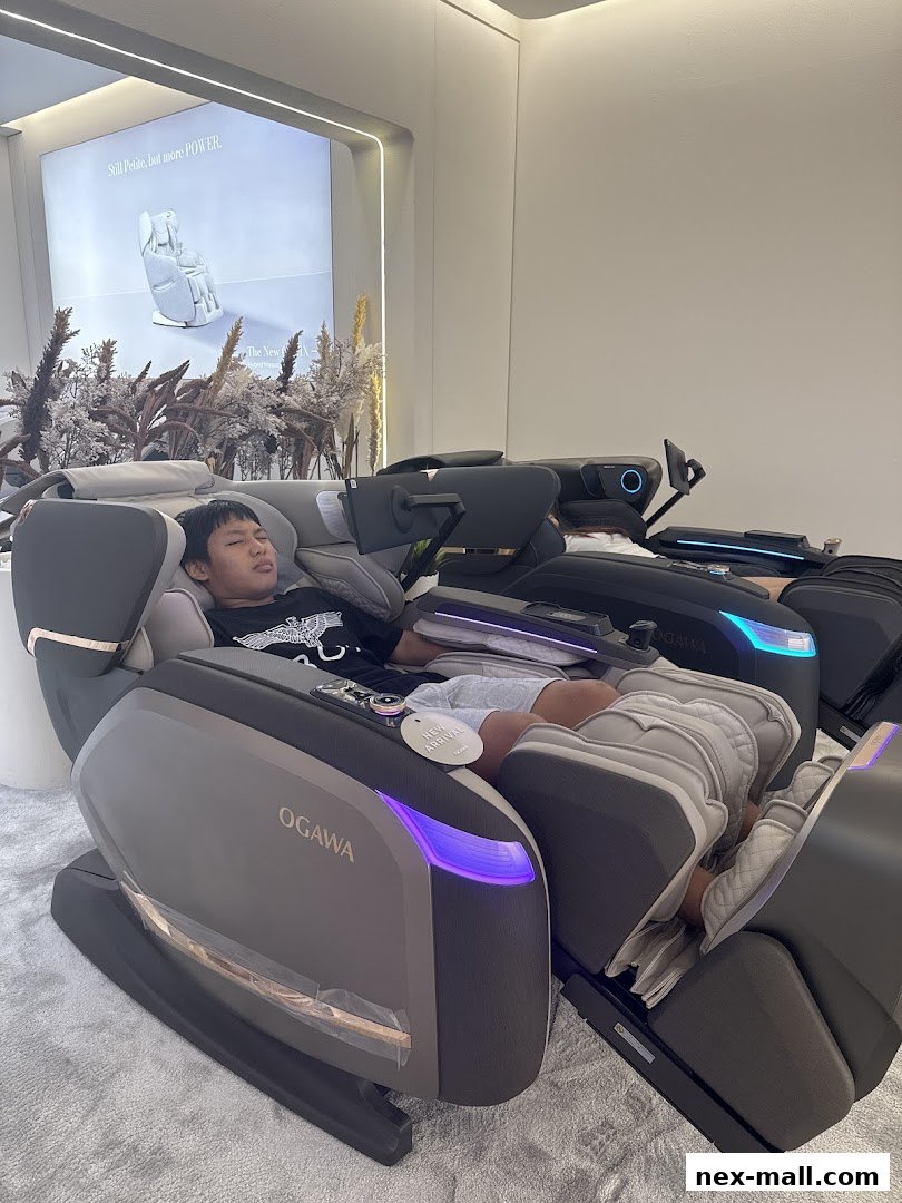 A person relaxing in an OGAWA massage chair with a promotional display in the background.