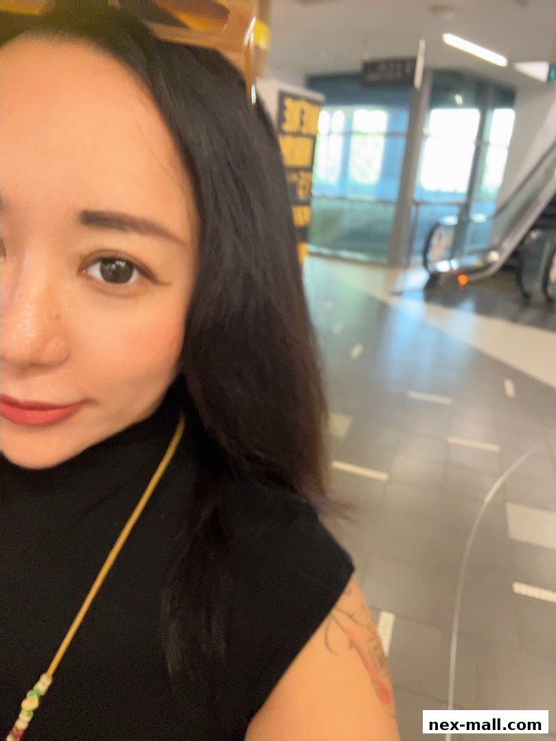 A woman with dark hair and makeup poses in a shopping mall with escalators in the background.
