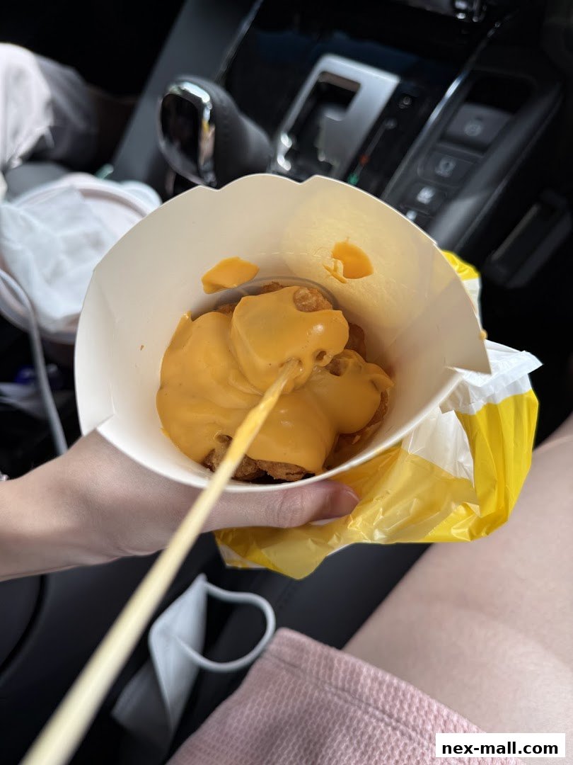 A hand holding a paper cup filled with fried snacks covered in cheese sauce.