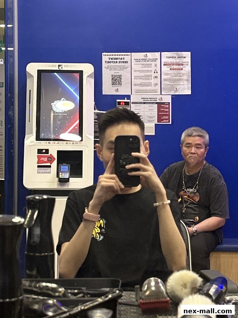 A person takes a selfie in a barber shop with a payment kiosk and posters on the wall.