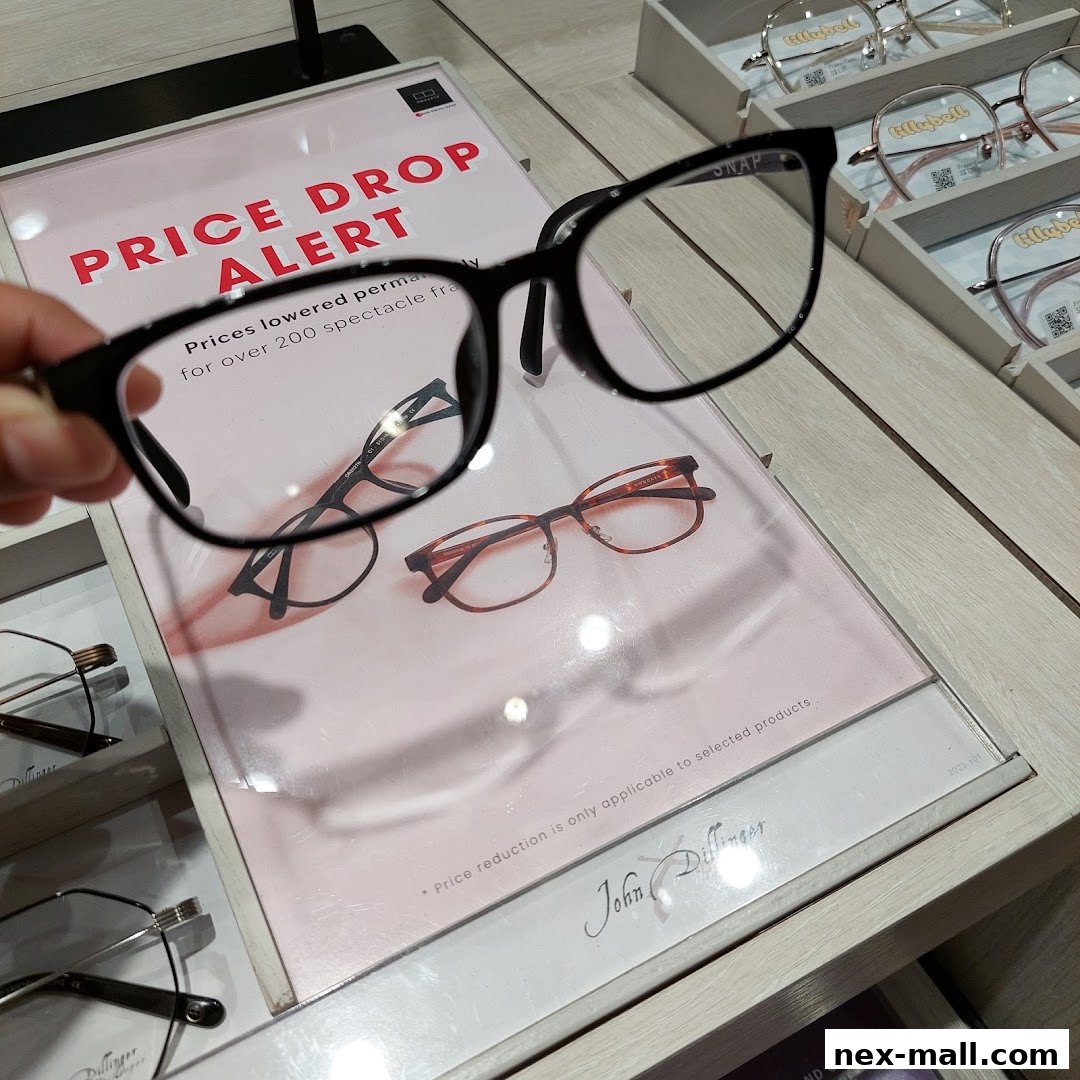 A hand holds up a pair of black-rimmed glasses in front of a display of eyeglasses.