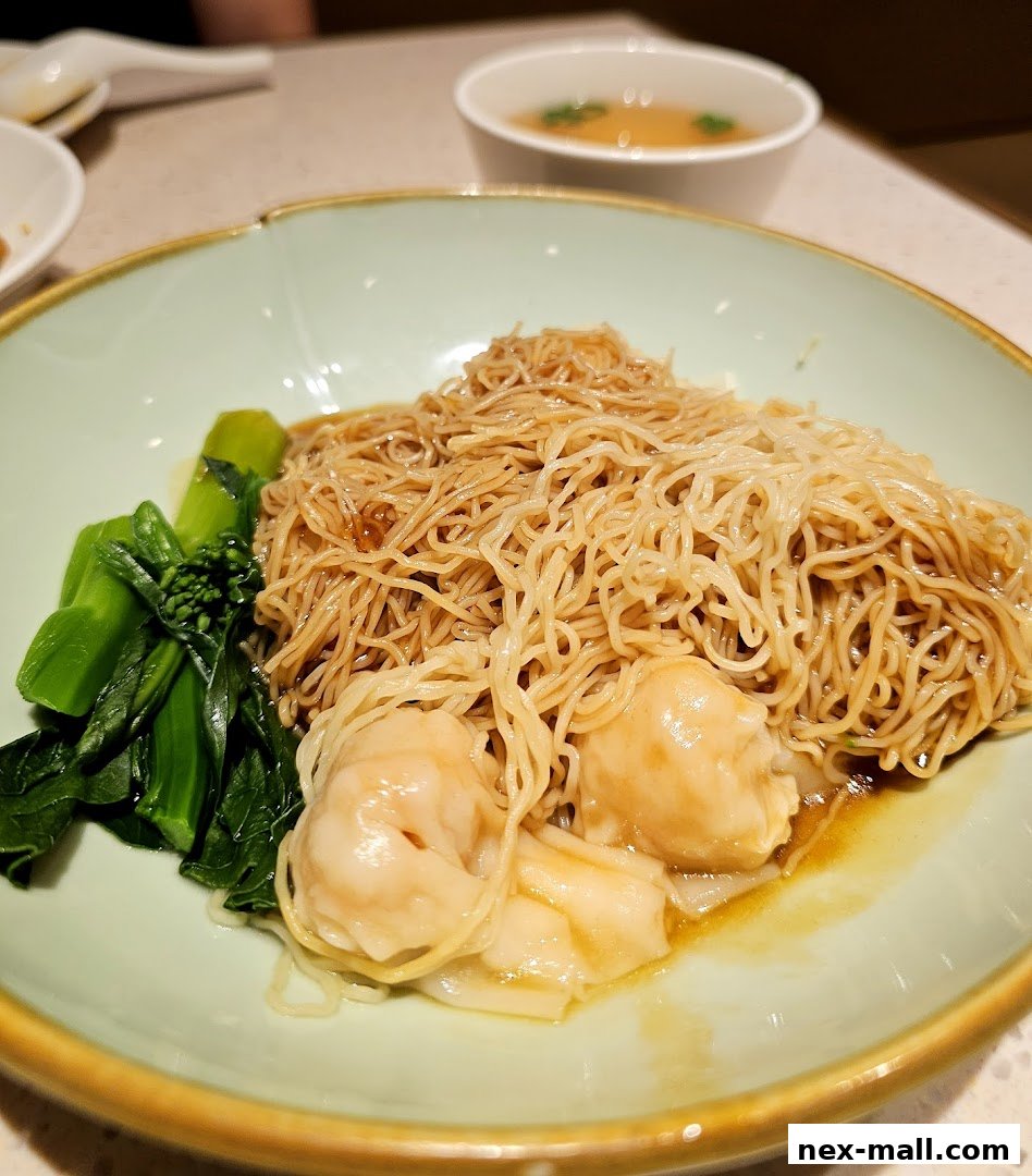 A close-up of a delicious wonton noodle dish with blanched greens and a side of broth.