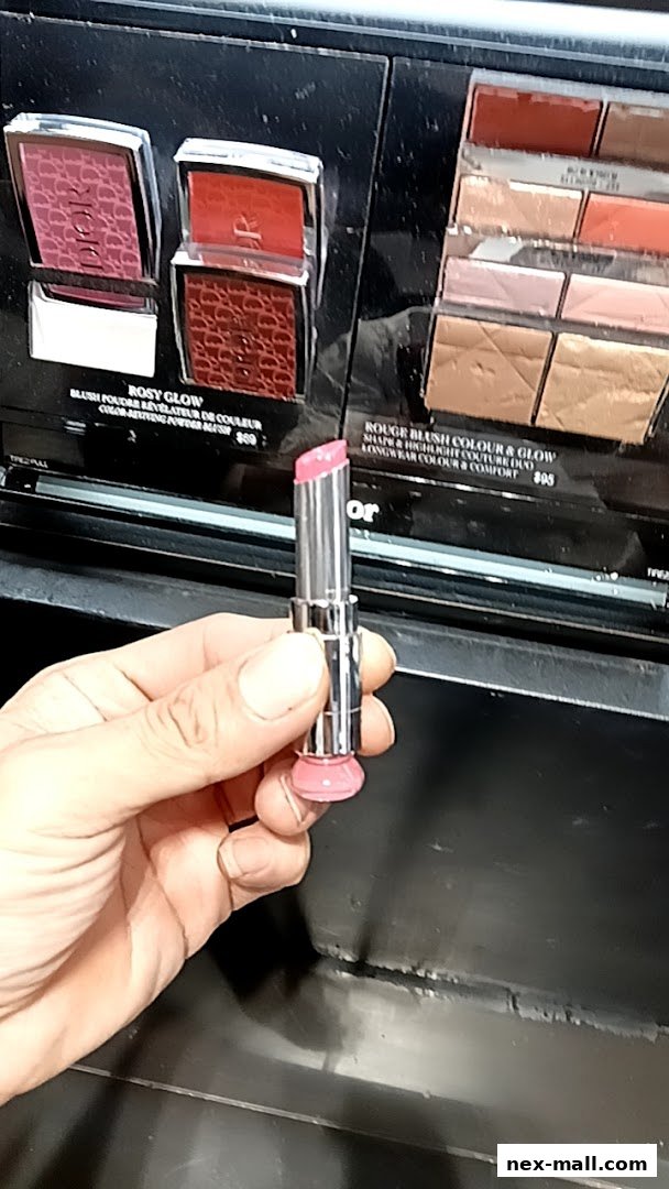 A hand holds a pink lipstick in front of a display of blushes and makeup palettes.