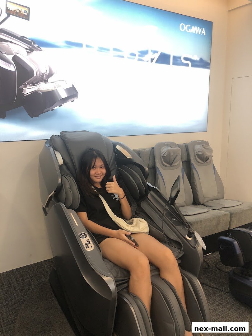 A young woman sits in a massage chair, giving a thumbs up, with other chairs in the background.