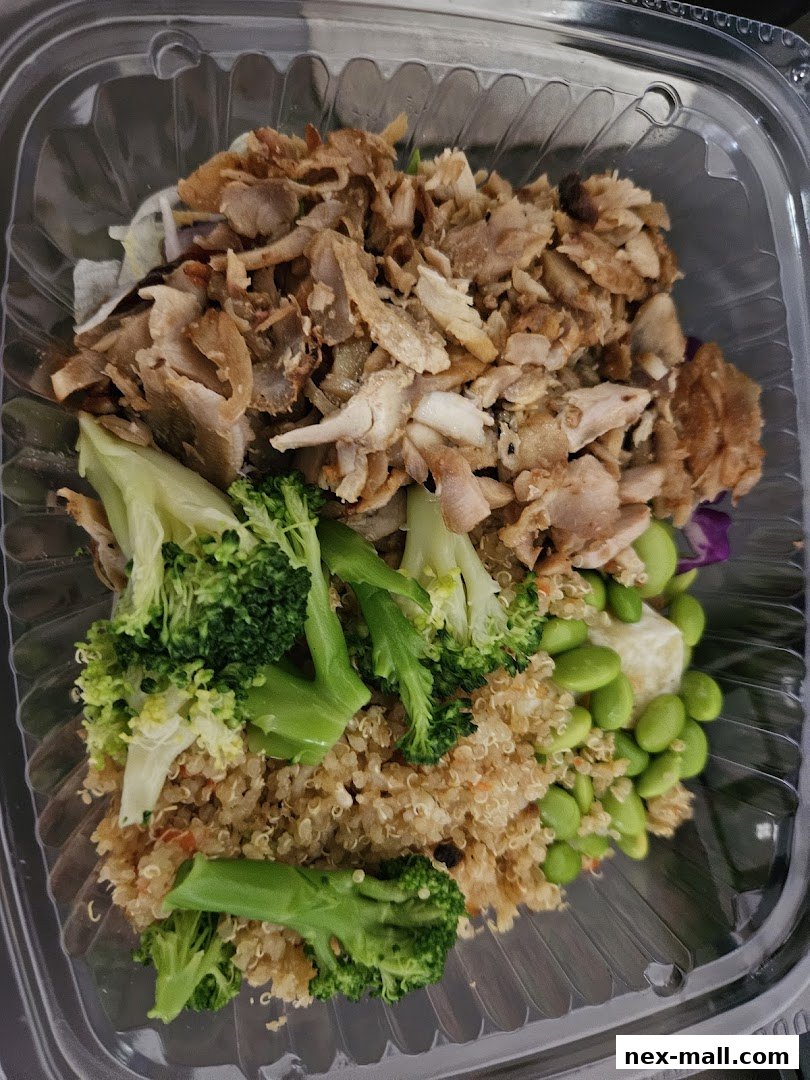 A clear plastic container filled with a healthy meal featuring grilled chicken, quinoa, broccoli, and edamame.