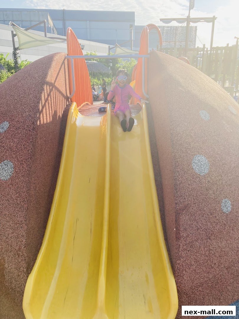 A young girl in a pink swimsuit and goggles slides down a yellow slide at a playground.