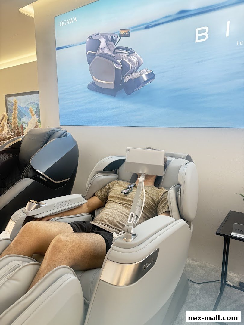 A man relaxes in a massage chair at the OGAWA NEX store, with a large advertisement behind him.