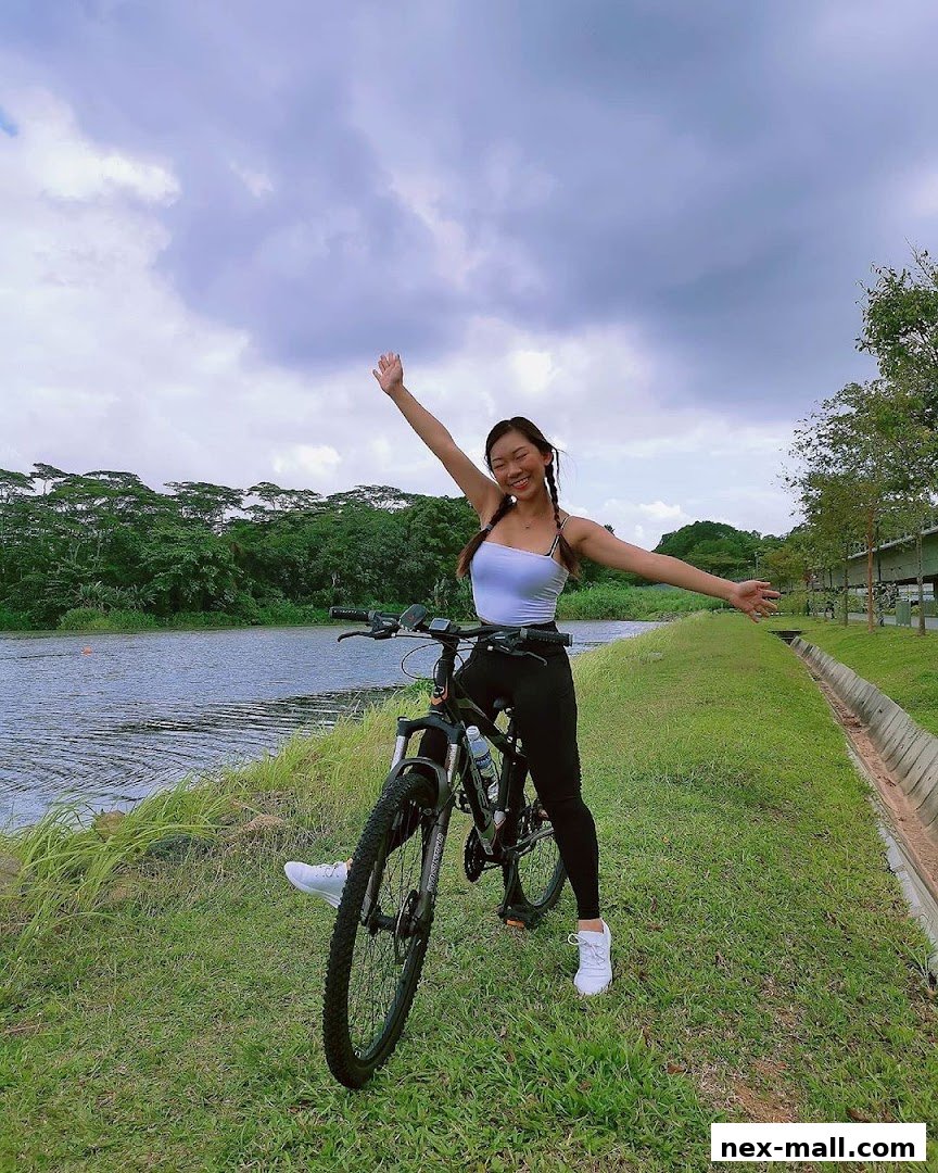 A woman with her arms outstretched poses with a bicycle by a lake.