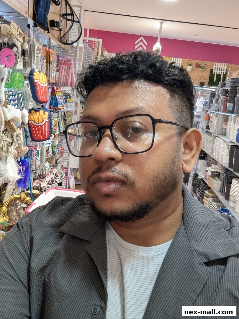 A man with glasses poses in front of shelves stocked with various items.