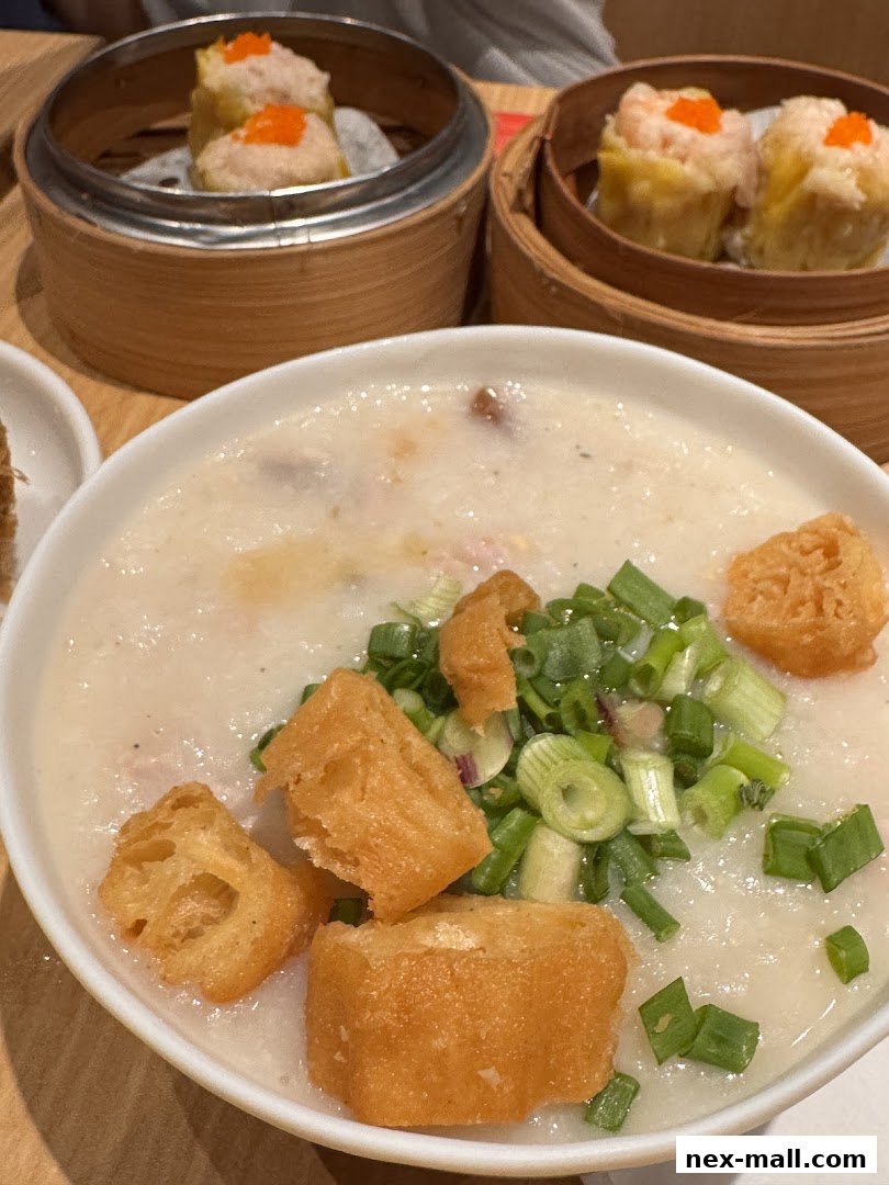 A close-up of a bowl of congee topped with scallions and fried dough sticks, with dim sum in the background.