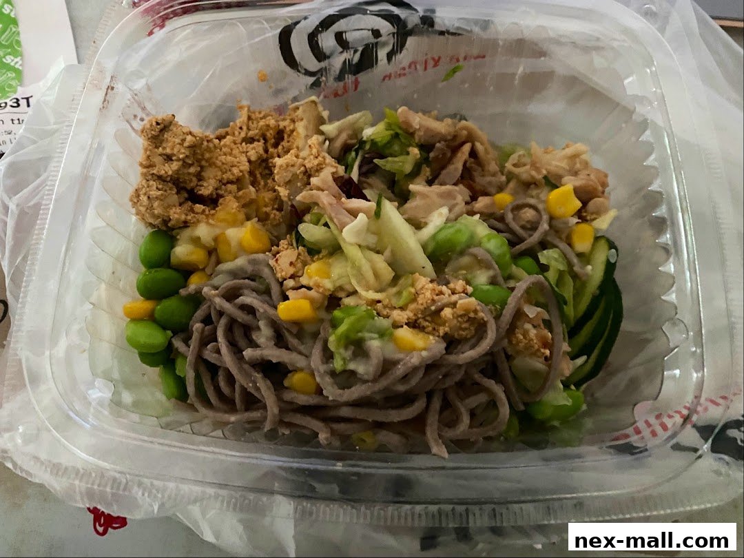 A clear plastic container filled with a healthy noodle salad with edamame, corn, and chicken.