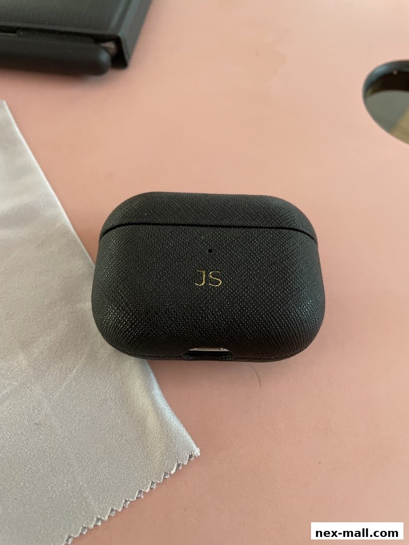 A personalized black AirPods case with gold initials 'JS' on a pink surface.