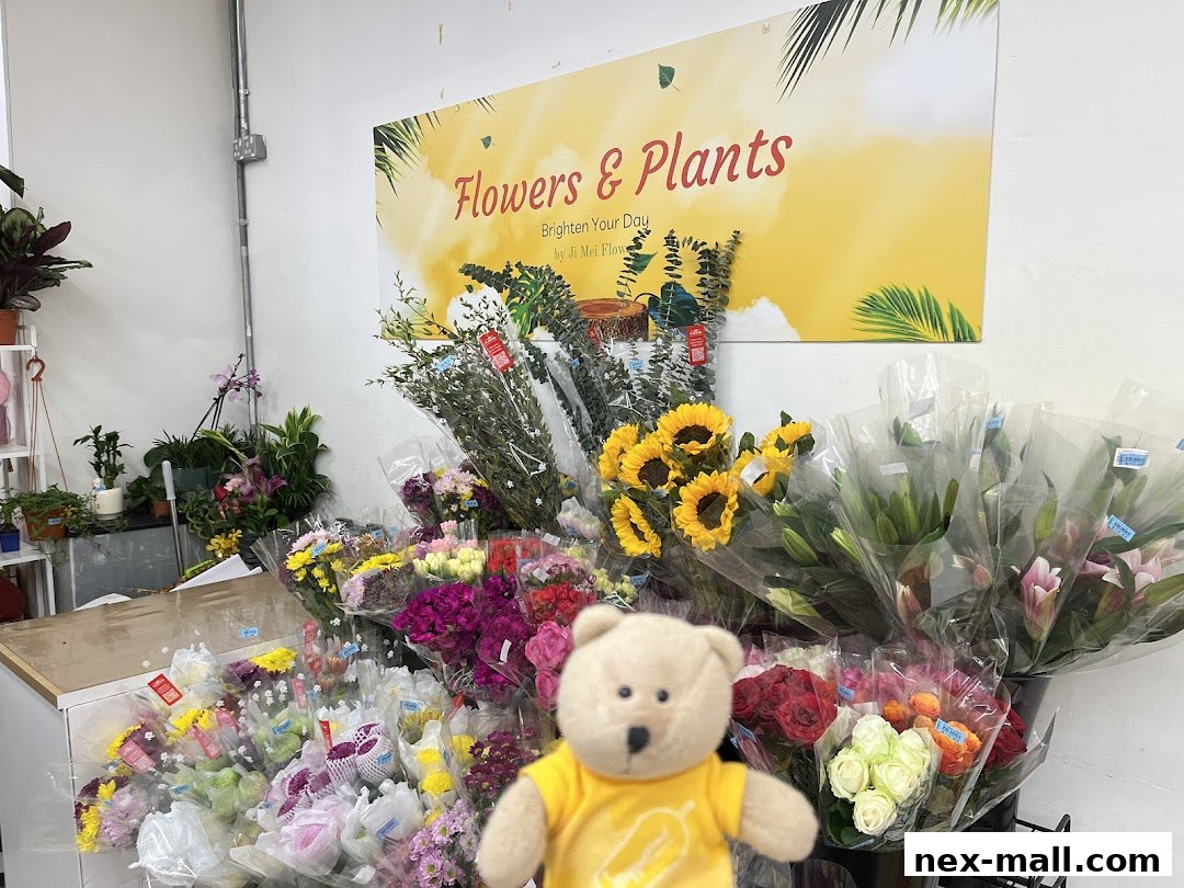 A variety of fresh flowers and potted plants are displayed in a shop, with a teddy bear in the foreground.