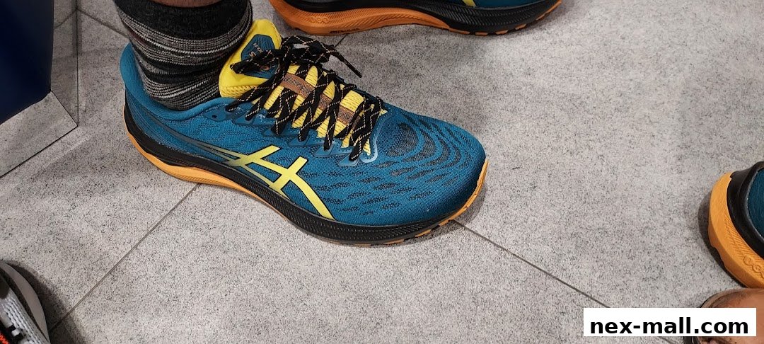 Close-up of a person wearing blue and yellow ASICS running shoes on a tiled floor.