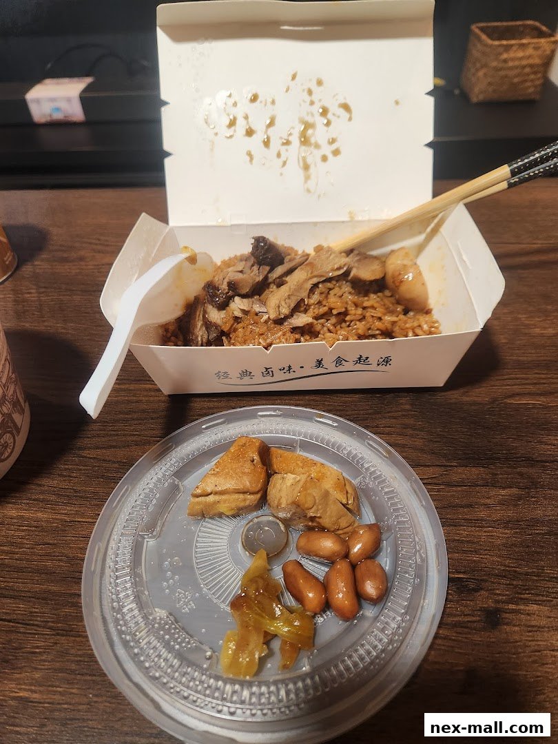 A takeout box filled with rice and meat, with a side of tofu, peanuts, and pickled vegetables.