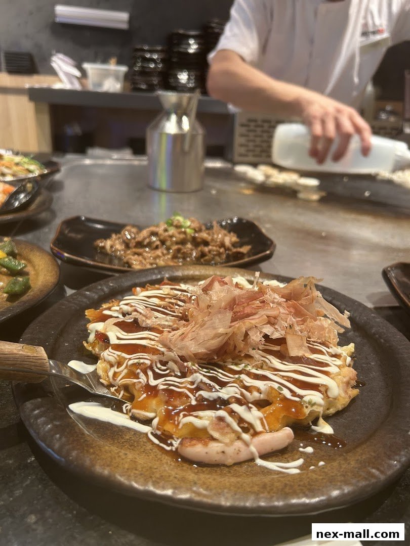 A close-up of a delicious okonomiyaki dish with a chef preparing food in the background.