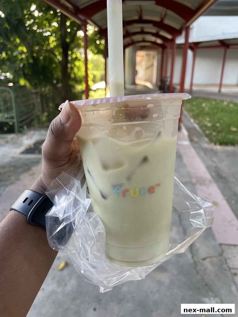 A hand holds a refreshing beverage from Fruce NEX, with a blurred background of an outdoor walkway.