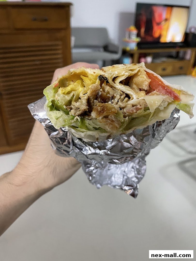 A hand holds a large, foil-wrapped wrap filled with meat, lettuce, and tomatoes.