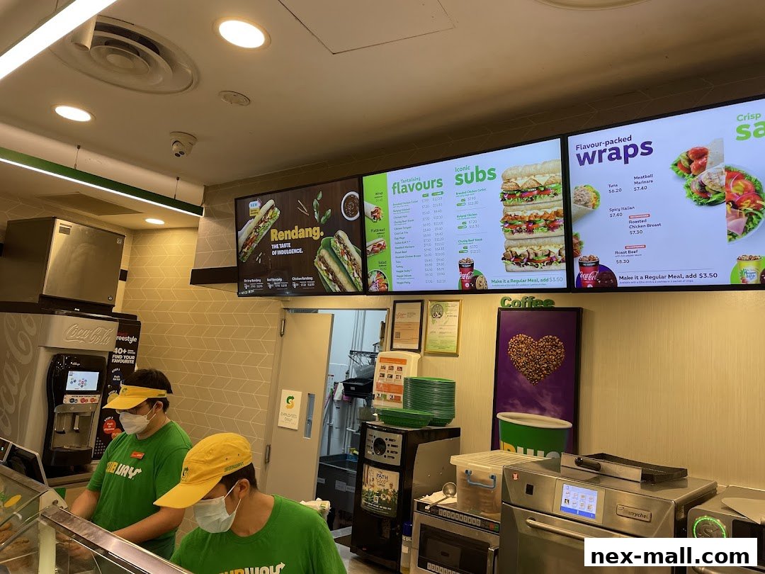 Subway employees prepare food behind a counter with digital menus displaying subs and wraps.