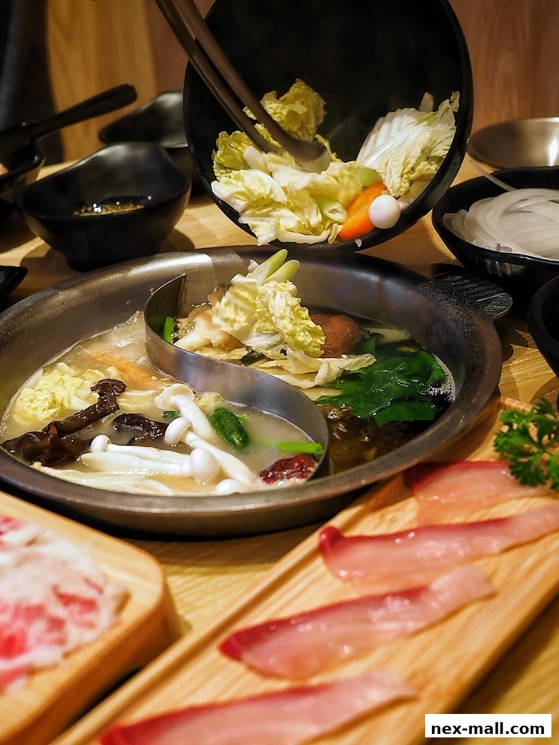 A split hot pot with various ingredients being added, including vegetables and thinly sliced meat.