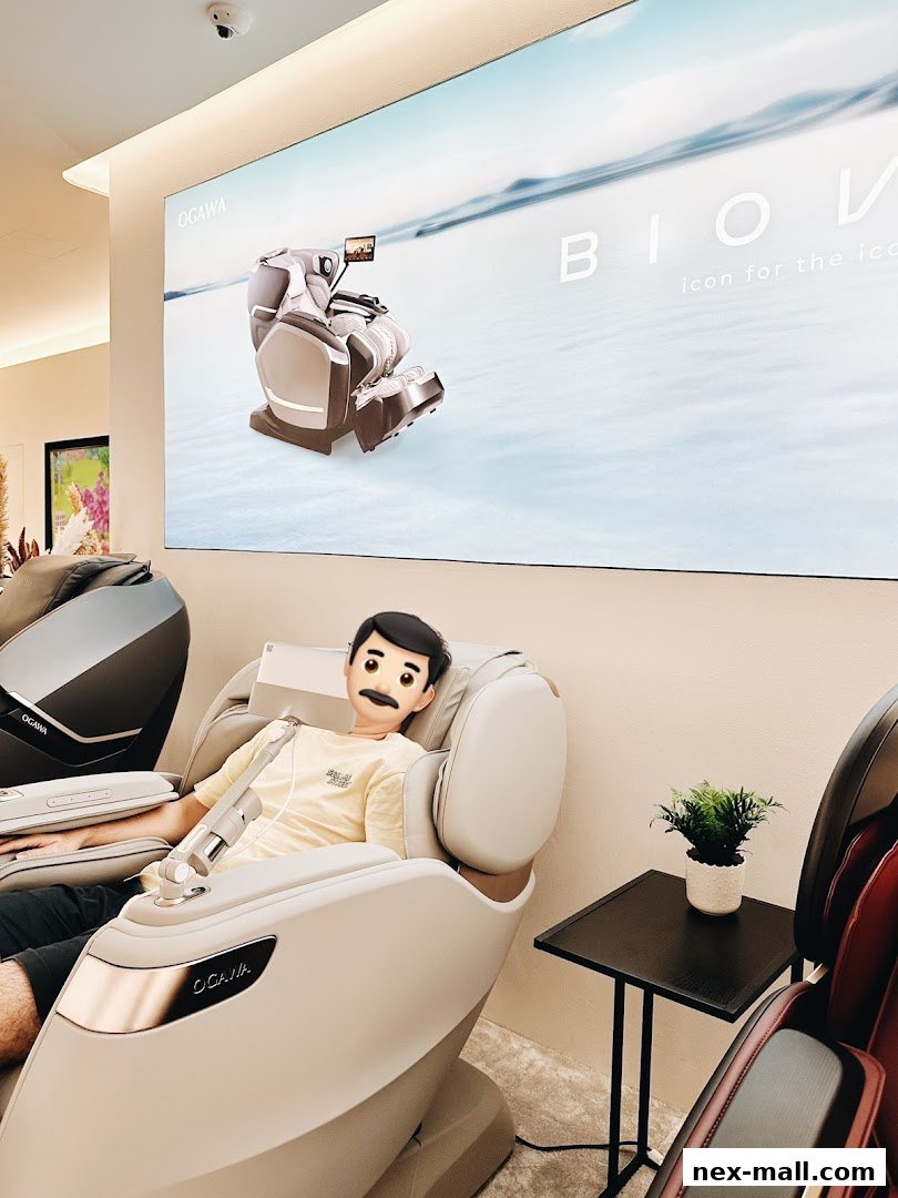 A person sits in a massage chair with a large screen displaying a massage chair and the OGAWA logo.