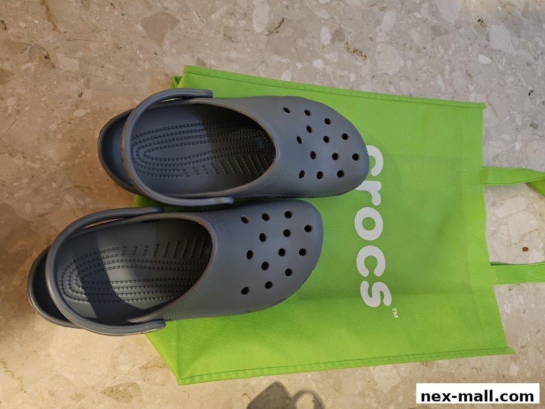 A pair of grey Crocs shoes resting on a bright green Crocs branded shopping bag.