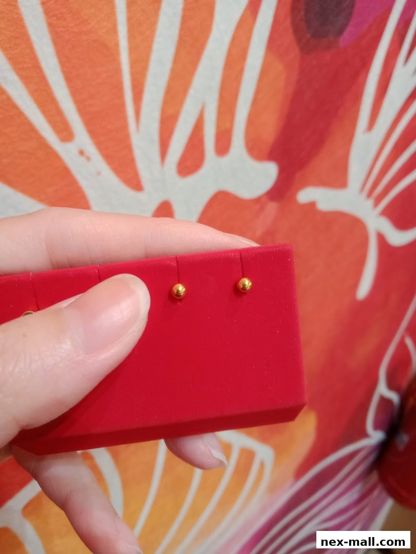 A hand holds a red jewelry box displaying small gold ball stud earrings.