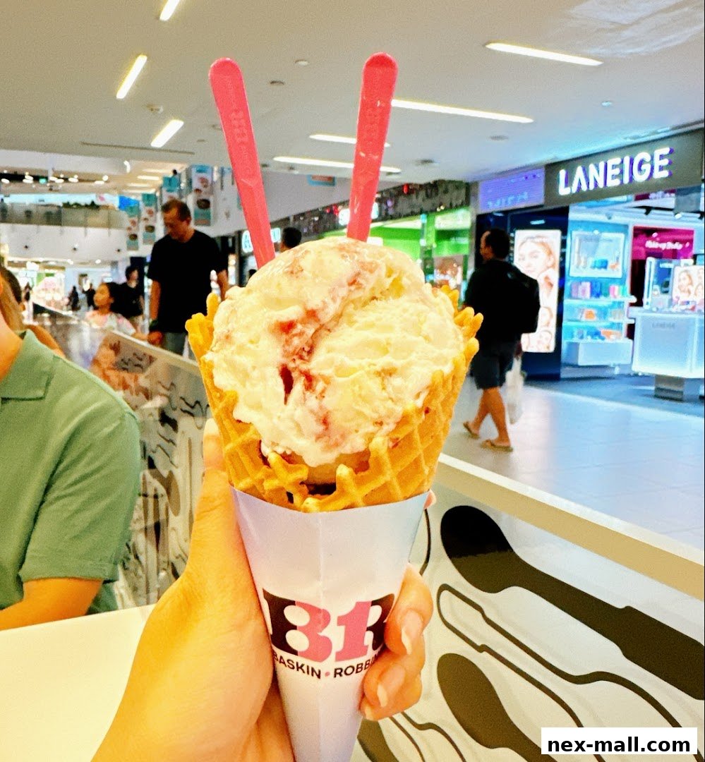 A hand holds a Baskin-Robbins ice cream cone with two pink spoons in a mall.