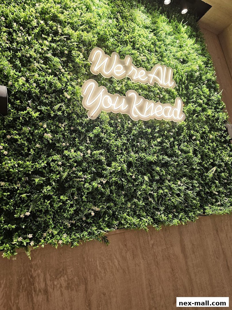 A neon sign saying 'We're All You Knead' on a lush green wall.