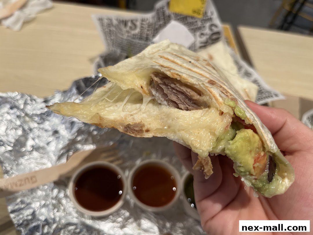 A hand holds a cheesy burrito filled with meat and avocado, with dipping sauces nearby.