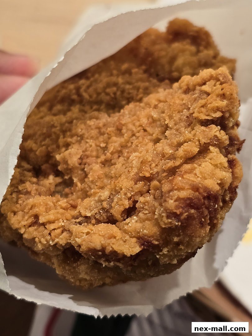 A close-up shot of a piece of crispy fried chicken held in white paper packaging.