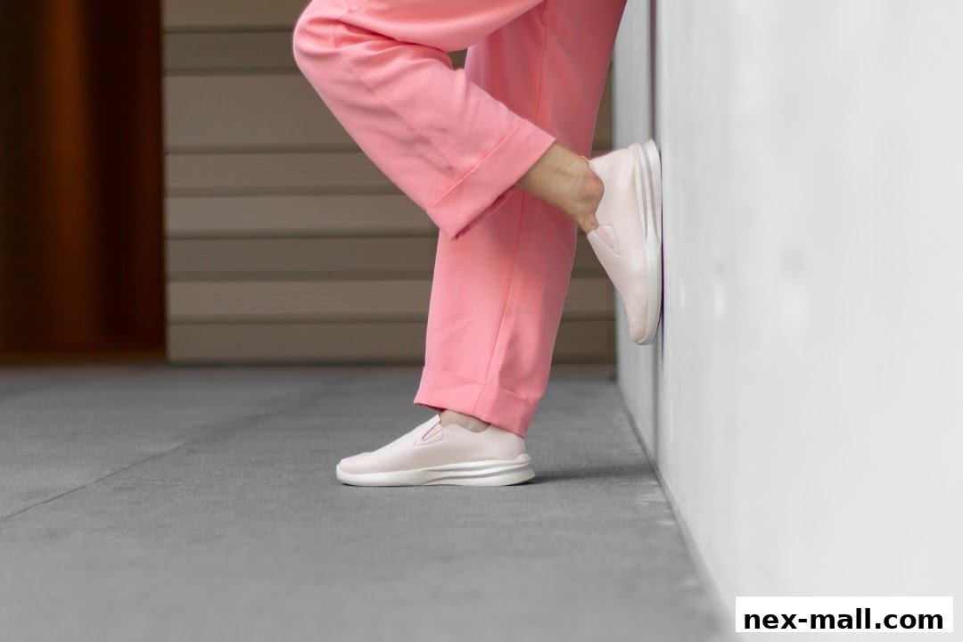 A person wearing pink pants and light pink slip-on shoes leans against a white wall.