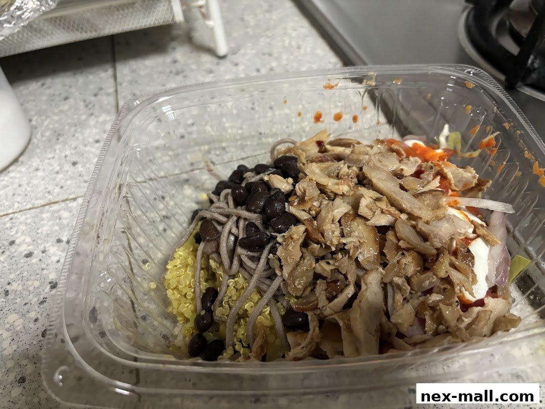 A clear plastic container filled with a healthy meal featuring quinoa, black beans, noodles, and shredded meat.