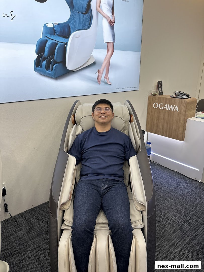 A man sits in a luxurious OGAWA massage chair, with a large advertisement for the chair in the background.