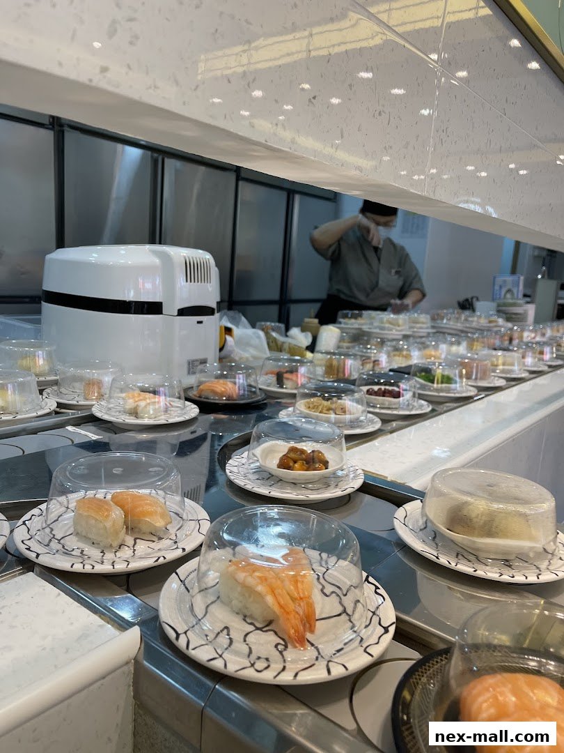 Sushi conveyor belt with various sushi dishes and a chef preparing food in the background.