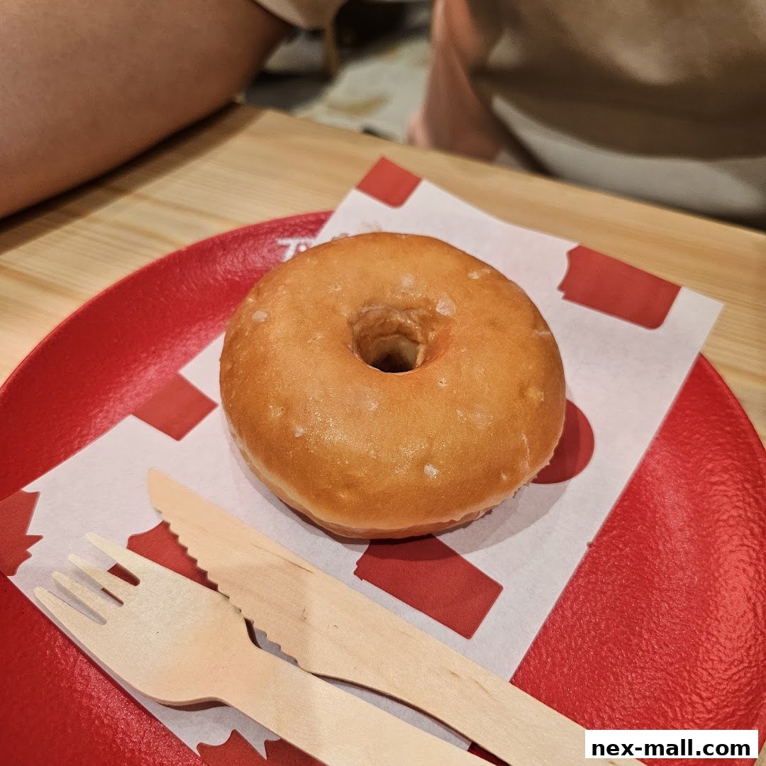 A glazed donut is served on a red plate with wooden cutlery and branded paper.