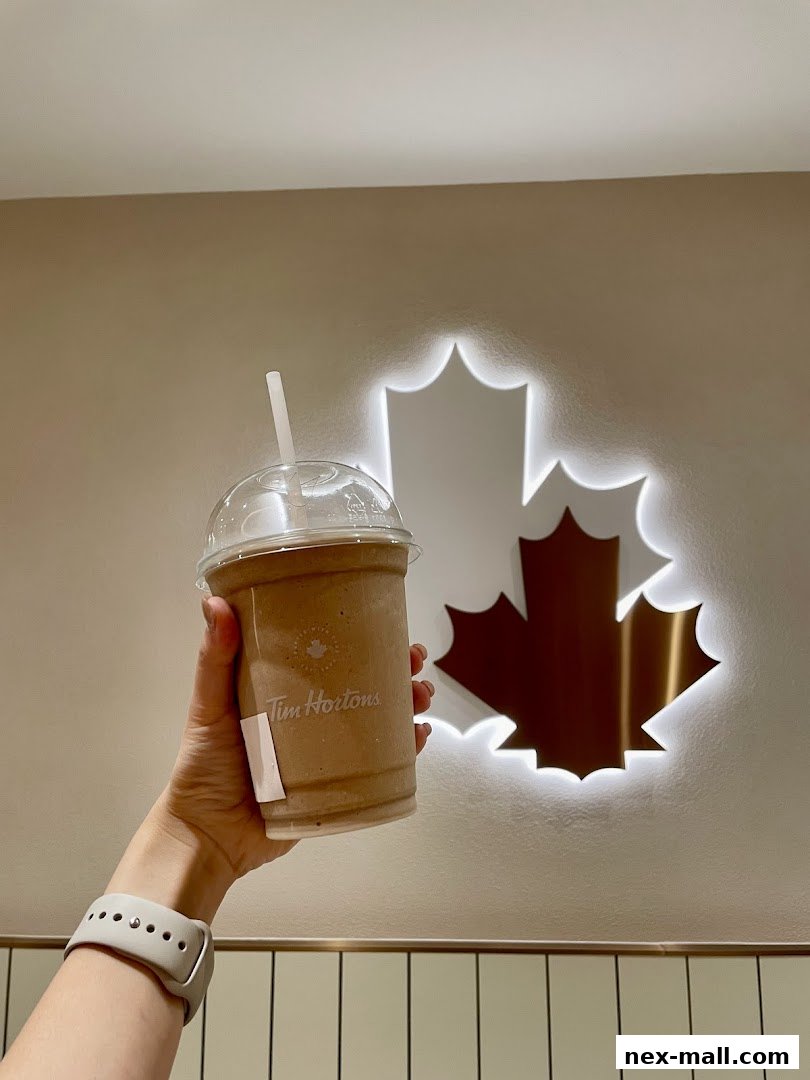 A hand holds a Tim Hortons iced coffee in front of a glowing Canadian maple leaf logo.