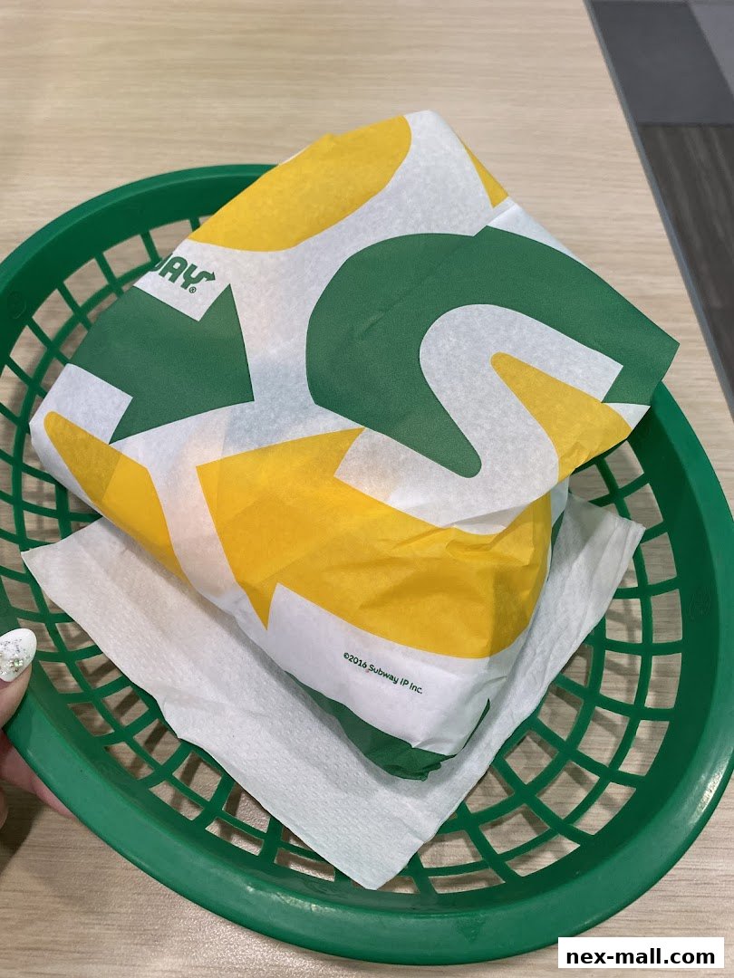 A Subway sandwich wrapped in branded paper, served in a green plastic basket.