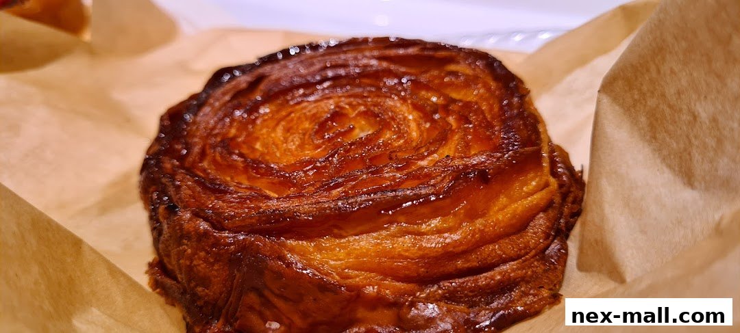 A close-up shot of a golden-brown, flaky pastry, likely a croissant or danish, wrapped in brown paper.
