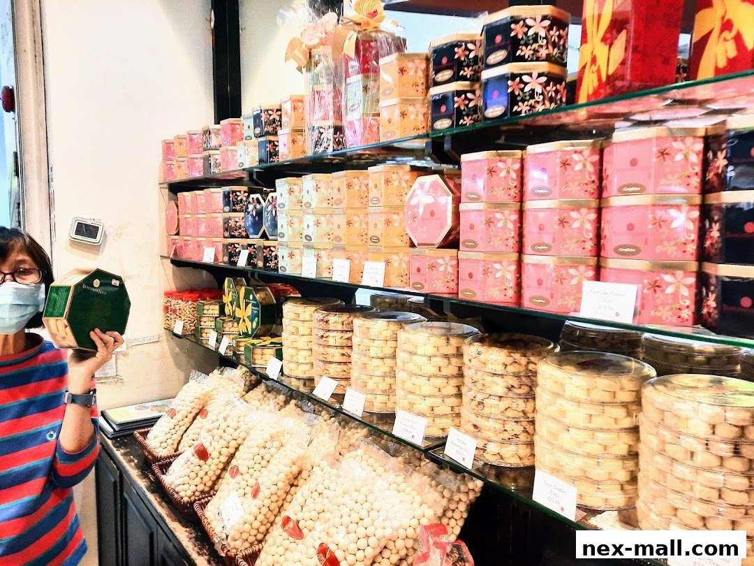 A woman in a mask holds a green box in front of shelves filled with colorful cookie tins and packaged cookies.