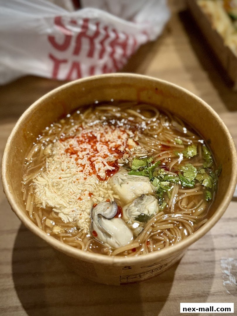 A close-up of a bowl of oyster mee sua with toppings and a blurred bag in the background.