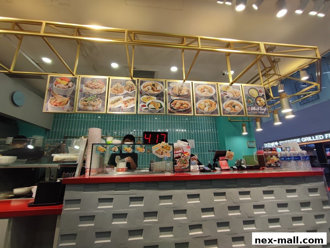 A food stall counter with a menu board displaying various dishes and a staff member serving customers.