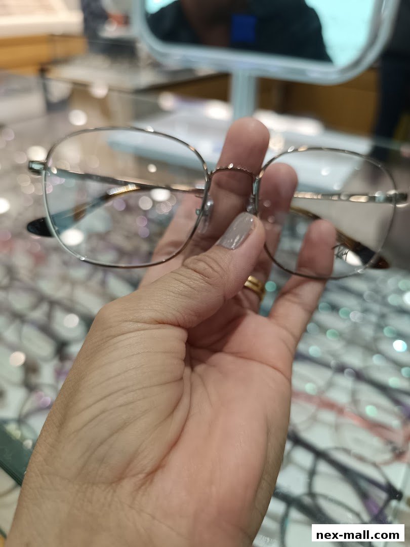 A hand holds a pair of stylish eyeglasses in front of a display of various eyewear.