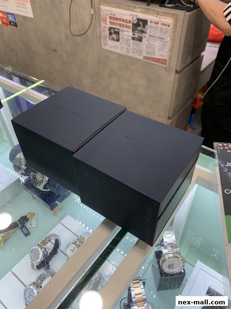 Two black boxes with 'ARIESGOLD' branding sit on a display case filled with watches.