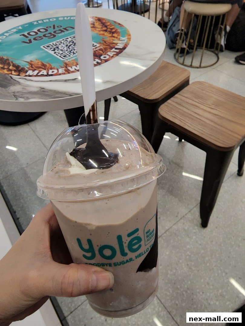 A hand holds a chocolate milkshake from Yolé Nex, with a table and stools in the background.