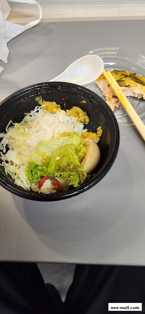 A close-up of a takeaway sushi bowl with rice, vegetables, and a hard-boiled egg, next to chopsticks and salmon.