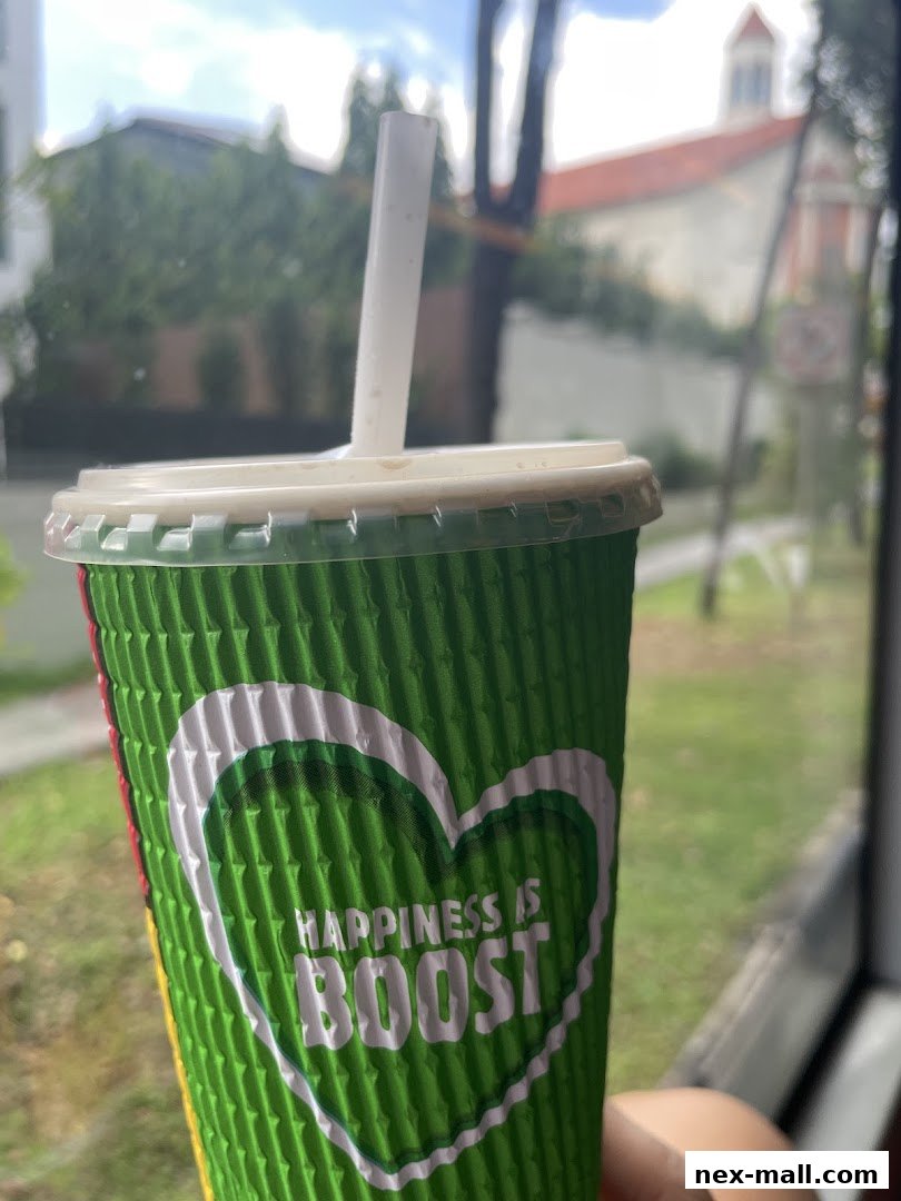 A green cup with a white straw and the Boost logo, held up against a blurred outdoor background.