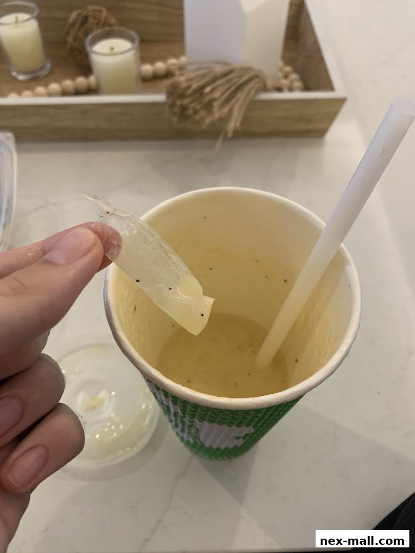A hand holds a piece of plastic film from a cup of smoothie with a straw.