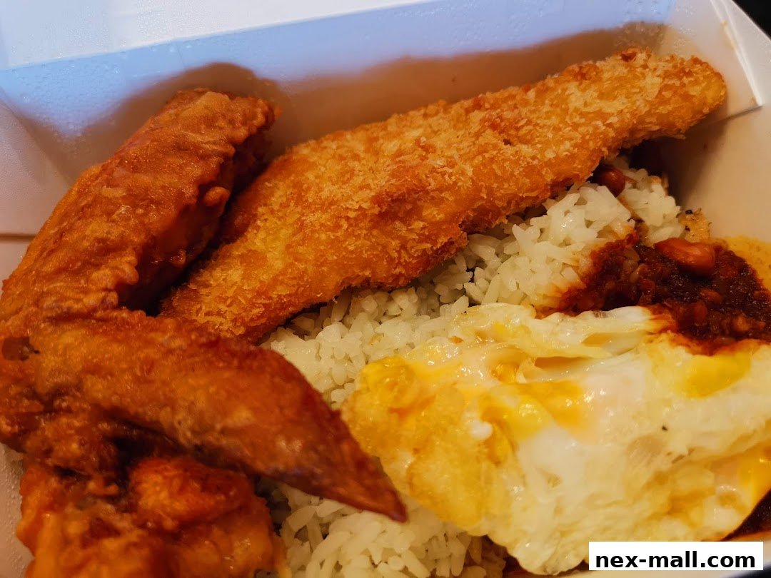 A close-up of a takeaway box filled with rice, fried chicken wing, fried fish fillet, fried egg, and sambal.