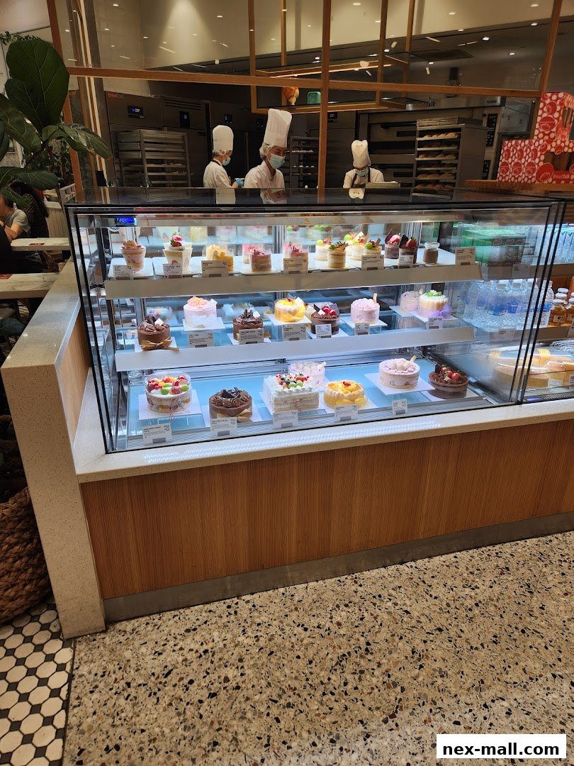 A display case filled with various cakes and pastries, with chefs visible in the background.